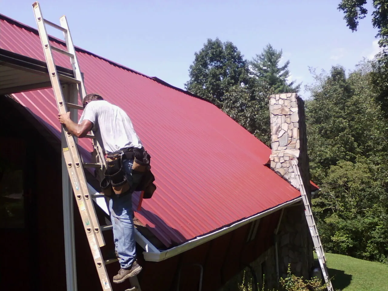 Roofer on a ladder beside a red metal roof and stone chimney — Soffit & Fascia Repair by Roofing Brothers in Carpentersville